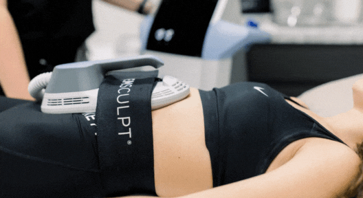 A woman lies comfortably while the Emsculpt device handpiece rests on her abdomen, delivering non-invasive muscle-toning treatment through gentle contractions.